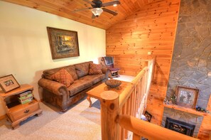 Una sala de estar acogedora con techo de madera, chimenea de piedra, sofá con cojines estampados y un cuadro en la pared.