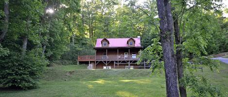 A cabin with a red roof, a porch, and a deck. It is surrounded by trees and a grassy area.