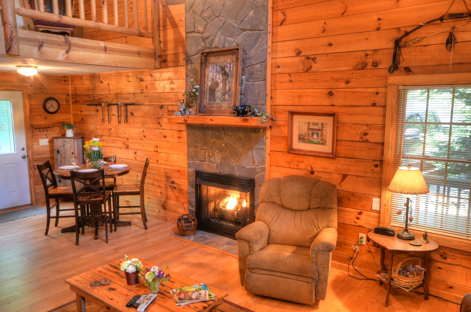 A cozy cabin interior with a stone fireplace, wooden walls, a dining area, and a comfortable armchair.