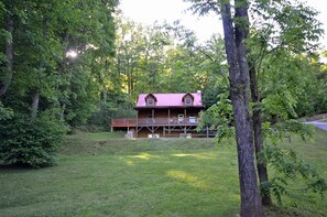 A cabin with a porch and a deck, surrounded by trees and a grassy area. - 2BR cabin with hot tub, game room, Wi-Fi, and mountain views. Free train and rafting tix (Whittier)