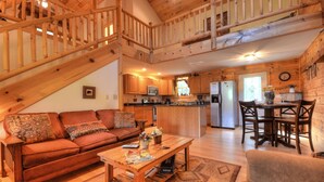A cozy cabin interior with a wooden staircase, a living room with a sofa and coffee table, a kitchen with wooden cabinets and a refrigerator, and a dining area with chairs.