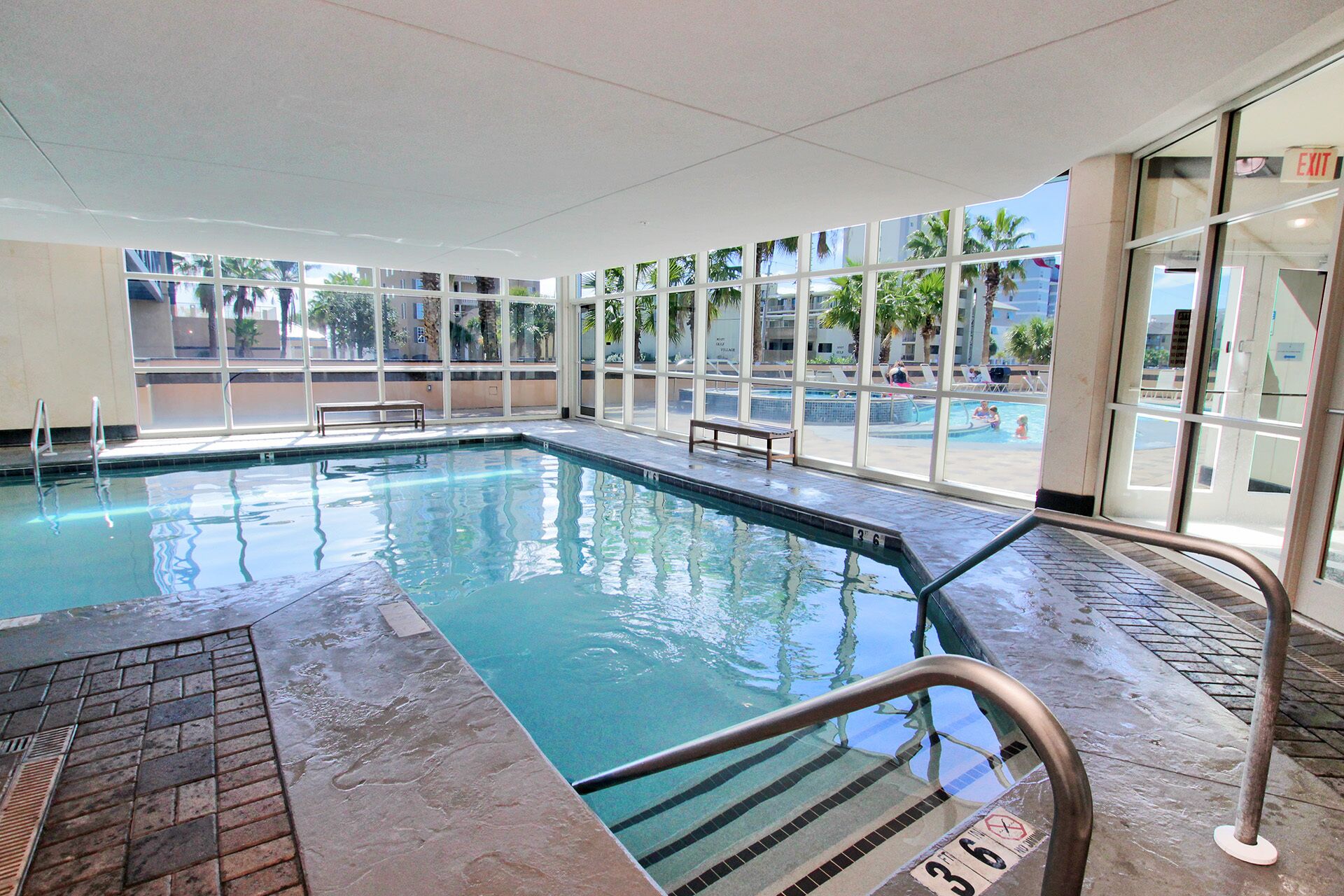 An indoor swimming pool with clear glass walls, a tiled floor, and a bench by the pool.