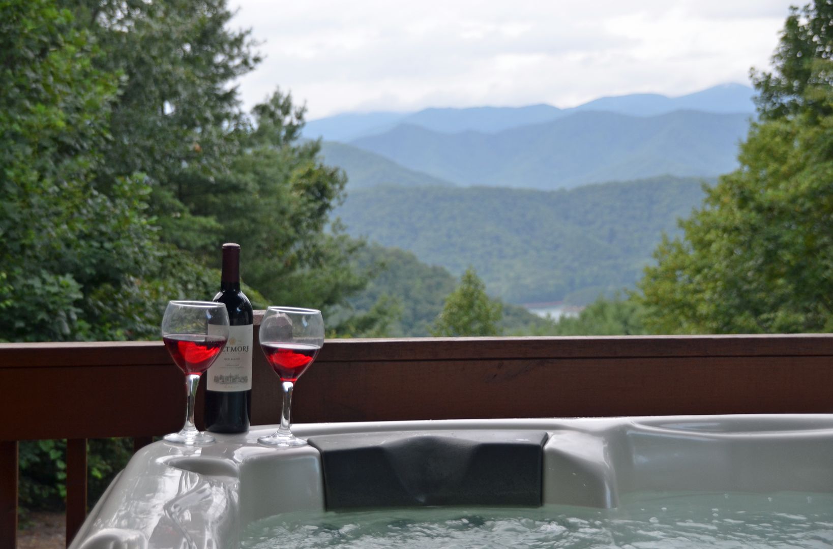 Un jacuzzi con dos copas de vino tinto y una botella, con un paisaje montañoso de fondo.