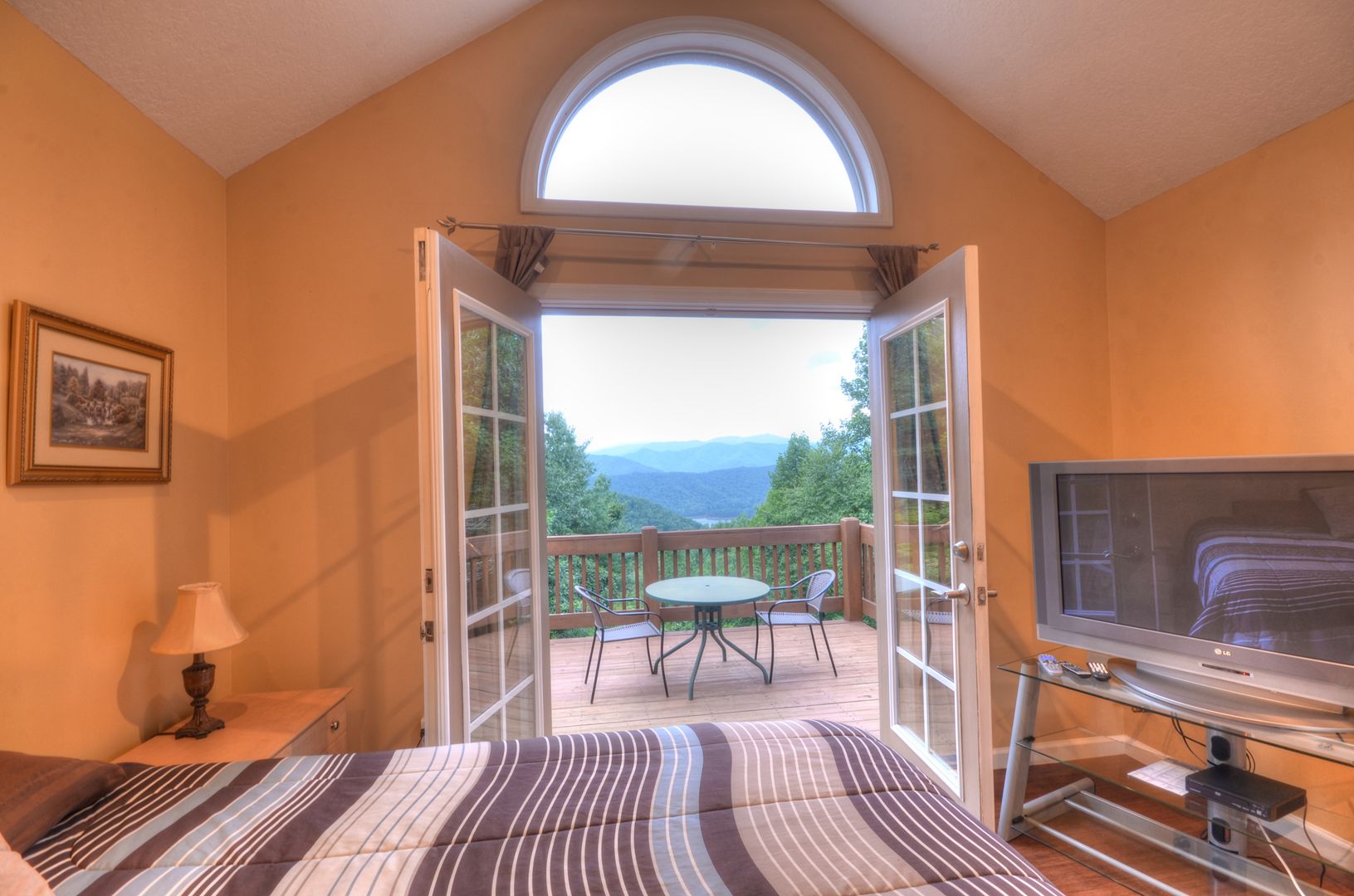 A bedroom with a large window, a bed, a TV, and a balcony with a table and chairs.