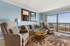 Ein Wohnzimmer mit einer Couch, Sesseln, einem Glastisch und Blick auf den Strand durch große Fenster.