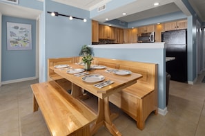 Uma área de refeições com uma mesa e bancos de madeira, uma cozinha com armários e eletrodomésticos de madeira, e um chão de azulejos.