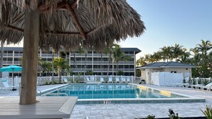A swimming pool area with a thatched umbrella, lounge chairs, and a building with multiple balconies in the background. - TRENDY BEACH CONDO ON THE GROUND FLOOR! (Sanibel)