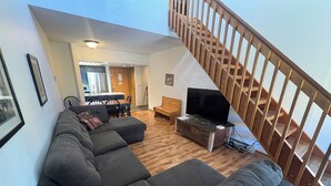 Ein Wohnzimmer mit Ecksofa, einem Fernseher auf einem Holzboden, einem Essbereich mit Tisch und Stühlen sowie einer Holztreppe.