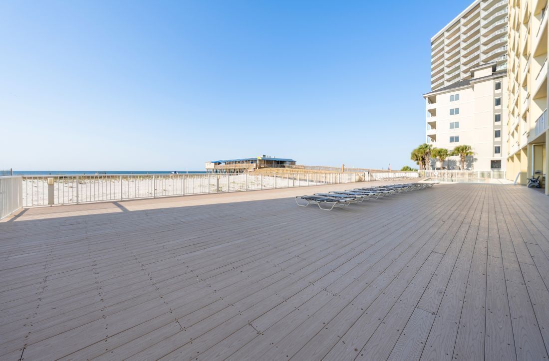 Un muelle de madera que conduce a una playa, con un edificio y palmeras al fondo.