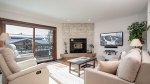 A modern living room with a stone fireplace, a television mounted on the wall, and a large window overlooking a snowy landscape. - RA215 | Mountain Setting | Excellent Amenities | Winter Shuttle | Wood FP (Steamboat Springs)