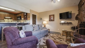 A living room with a sofa, two armchairs, a television, a fireplace, and a kitchen in the background. - SM323 | Slopeside Condo | Hot Tub | Ski In/Walk Out | Free City Bus (Steamboat Springs)