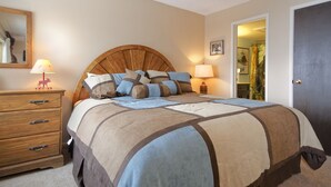 A bedroom with a wooden headboard, a bed with a patterned bedspread, two bedside lamps, a wooden dresser with drawers, and a door leading to another room. - SM323 | Slopeside Condo | Hot Tub | Ski In/Walk Out | Free City Bus (Steamboat Springs)