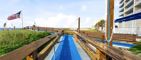 Une promenade en bois menant à une plage où flotte un drapeau américain, avec un chemin bleu et un bâtiment doté de balcons.