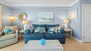 Un soggiorno con un divano blu, due tavolini con lampade, un tavolino da caffè con un vaso blu e un muro con un quadro.