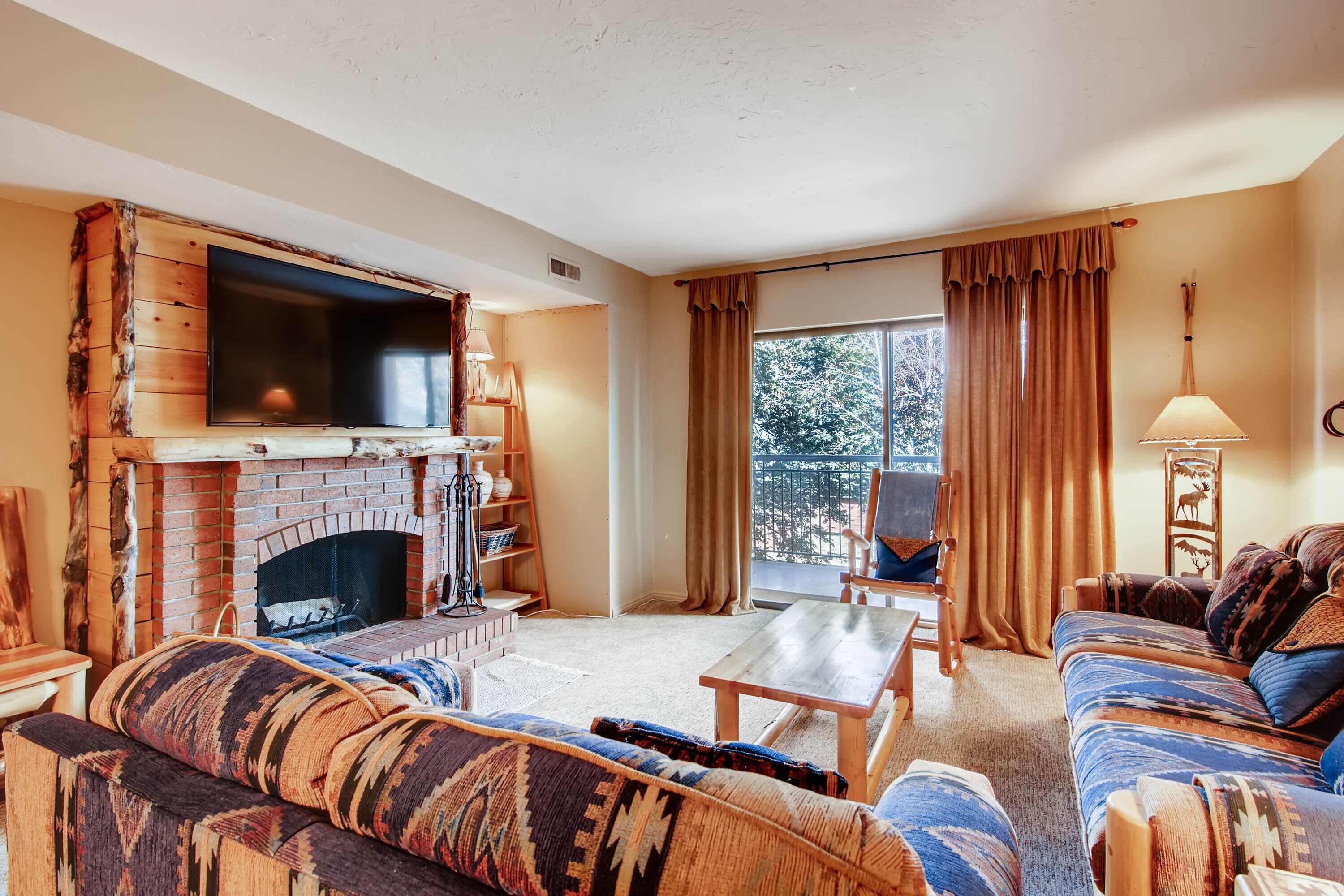 A cozy living room with a brick fireplace, a flat-screen TV, a sofa, a coffee table, and a view of trees through a large window.