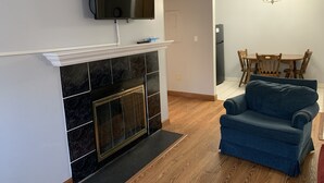 A living room with a fireplace, a flat-screen TV mounted above it, a blue sofa, and a dining area with wooden chairs and a table. - Oak Square 109 is a spacious condo with private balcony in downtown (Gatlinburg)