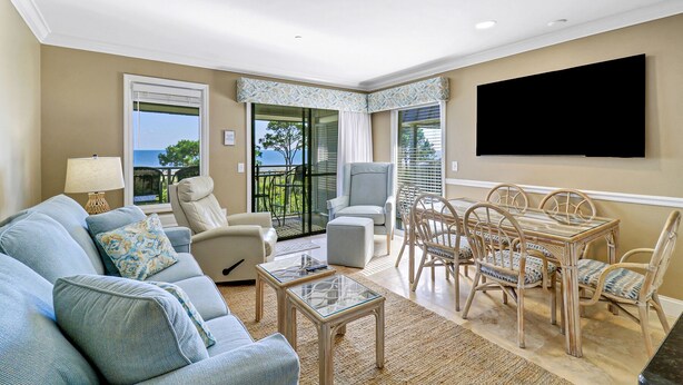 A living room with a sofa, armchairs, a glass coffee table, a dining table with chairs, a TV mounted on the wall, and a view of the sea through large windows. - Stunning Oceanfront Villa! Heated Beach Side Pool (Hilton Head)