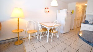 A small, clean kitchen with white cabinets, a white refrigerator, a wooden dining table with four chairs, a floor lamp, and a life preserver hanging on the wall.