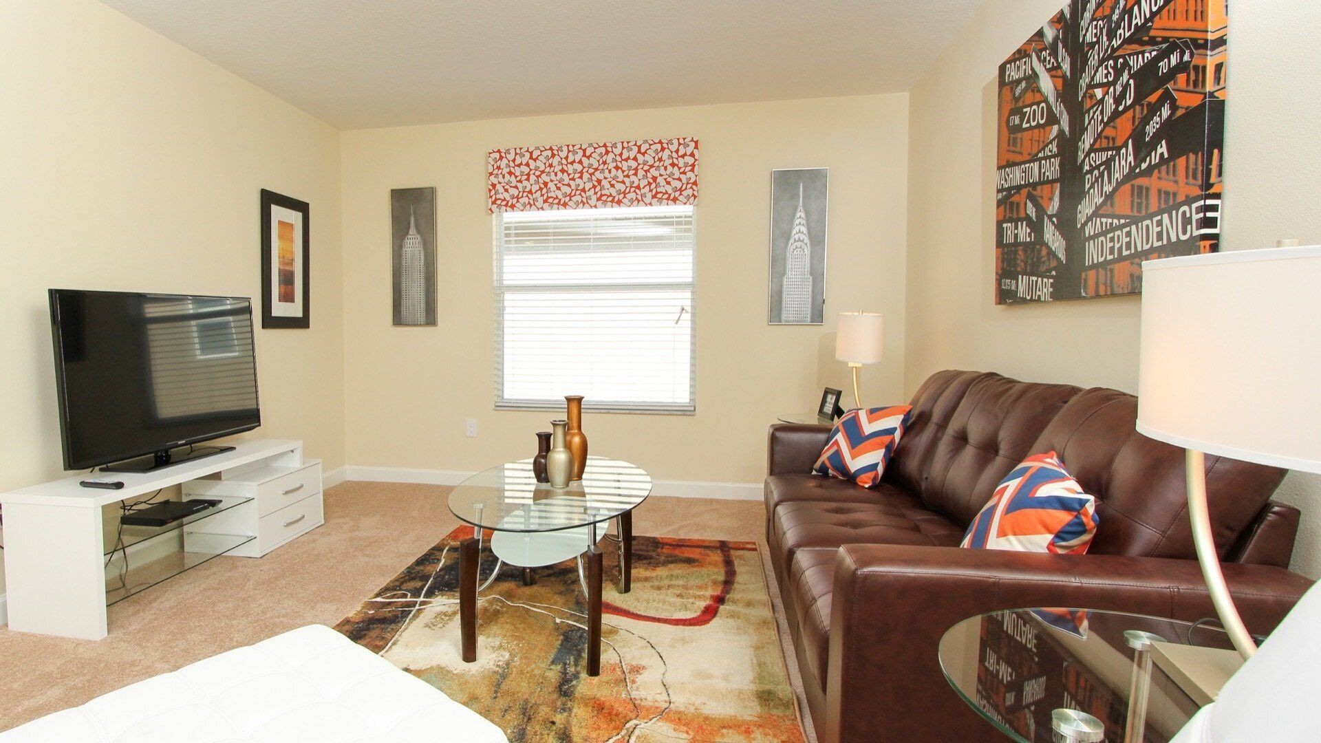 A living room with a flat-screen TV, a glass coffee table, a brown leather sofa, and a patterned rug.