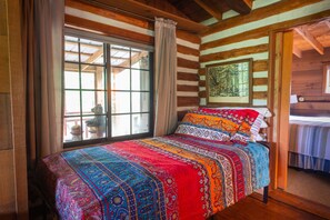 Una habitación tipo cabaña de madera con un ventanal grande, una cama con una manta colorida y un cuadro en la pared.