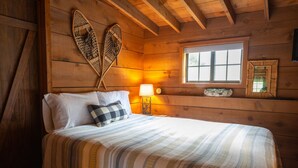 Habitación de cabaña de madera con cama, almohadas, una lámpara, una ventana y un espejo montado en la pared.