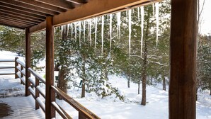 Un área cubierta de madera con barandal de madera, suelo cubierto de nieve y árboles al fondo.