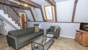 Et loft-agtigt opholdsrum med et sofabord i glas, to sofaer, et træskab og en trappe med synlige bjælker.