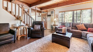 A spacious living room with a wooden ceiling, a leather sofa, a black armchair, and a staircase with wooden beams.