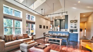A spacious living area with a sofa, ottoman, and dining table. A kitchen with blue cabinets and a large island is visible in the background.