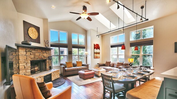 A spacious living room with a stone fireplace, leather sofas, a dining area with a wooden table, and large windows providing natural light.