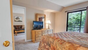 Un dormitorio con cama, cómoda, televisor y ventana con vista a árboles.