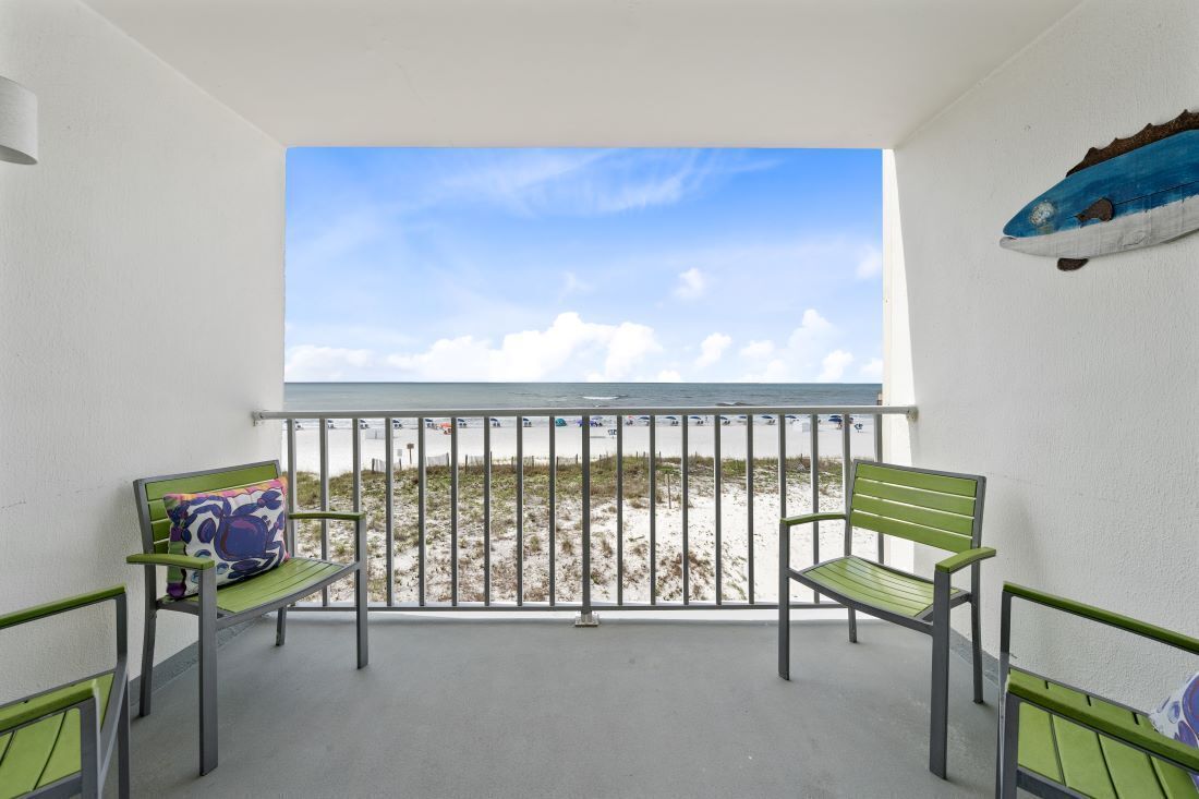 A balcony with two green chairs and a colorful cushion, overlooking a beach with a clear sky.