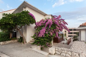 A building with a stone staircase leading to a door, surrounded by greenery and purple flowers.