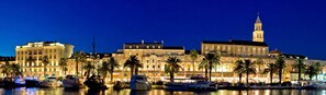 A waterfront hotel with a prominent building featuring a tower, surrounded by palm trees and boats.