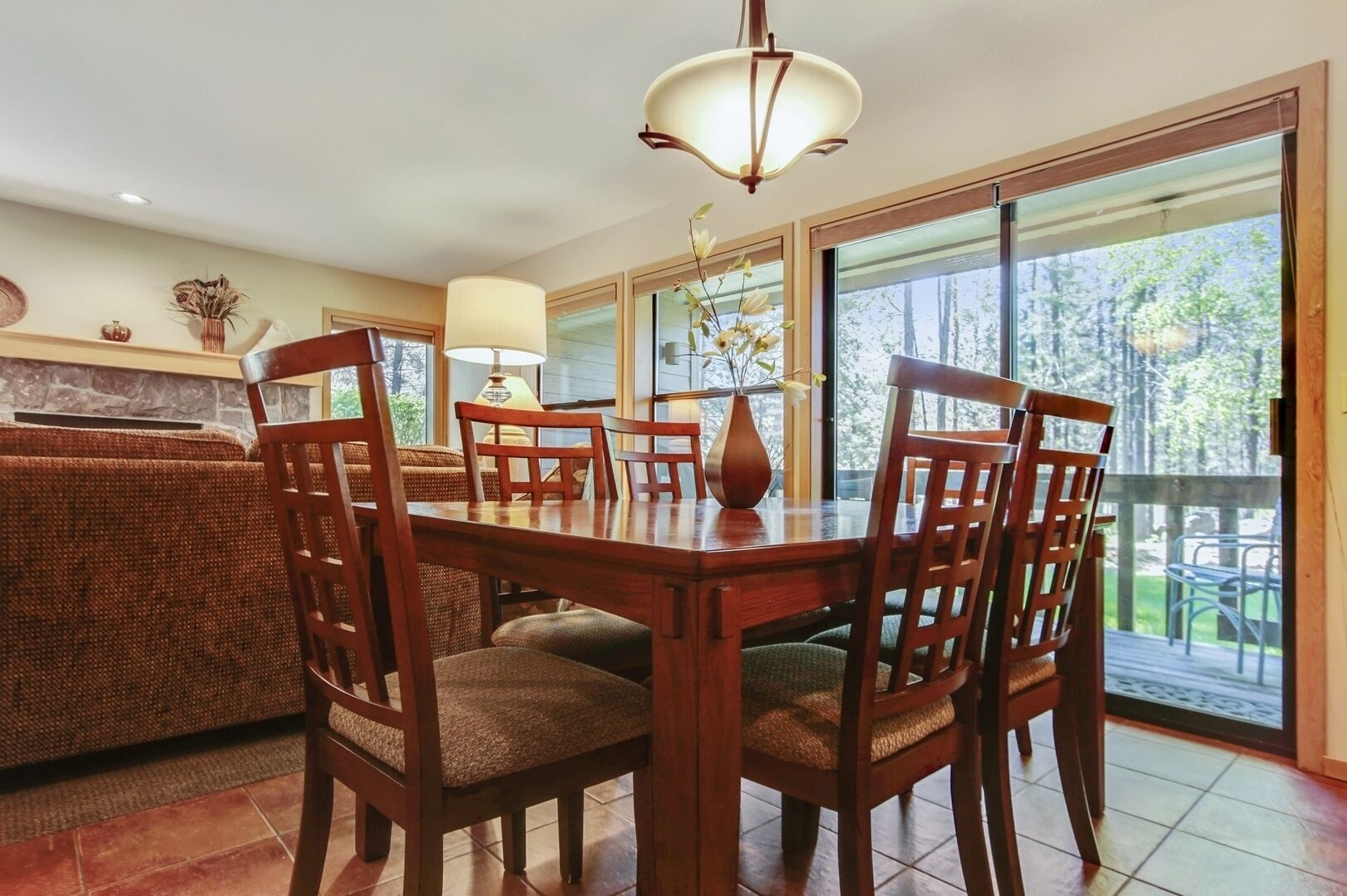 A dining area with a wooden table and chairs, a vase with flowers, and a view of the outdoors through large windows.