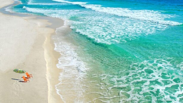 Ein Strand mit klarem türkisblauem Wasser, einem Sandstrand und einem Segelboot in der Ferne.