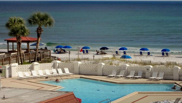 A pool area with lounge chairs and umbrellas, a beach in the background, and a clear view of the ocean.