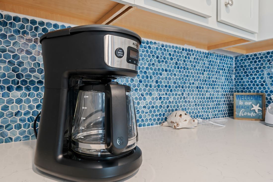 Una cafetera sobre una encimera de cocina con un salpicadero de azulejos hexagonales azules y un cartel que dice 'Welcome to our Beach Home'.