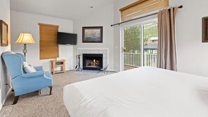 Ein Hotelzimmer mit einem großen Bett, einem blauen Sessel, einem Kamin, einem Fernseher und einem Balkon mit Blick nach außen.