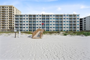 A beachfront hotel with multiple balconies, a sandy beach in front, and a wooden boardwalk leading to the entrance.