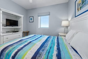 A bedroom with a bed, a TV on a shelf, a window with blinds, and a bedside table with a lamp.