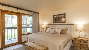 A bedroom with a large bed, two bedside tables with lamps, a wooden bench, and a sliding glass door leading to an outdoor area. - Hotel Style Room in the Heart of WMR Village (Whitefish)