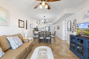 A spacious living room with a brown sofa, dining area with wooden chairs, a TV on a blue cabinet, and a ceiling fan.