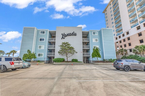 A hotel with a central sign reading "Vanaqua," featuring a modern design with light blue and beige colors, multiple floors, and a parking lot with several cars.