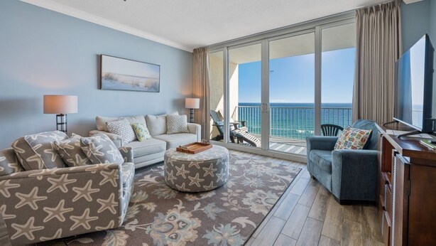 A living room with a sofa, ottoman, and a TV, with a balcony and ocean view. - Beachfront w/ Expansive Gulf Front View | Pool, Outdoor Grills, Sauna | Sleeps 7 (Gulf Shores)