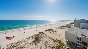 A beach view with a flag, clear sky, and buildings in the background. - Beachfront w/ Expansive Gulf Front View | Pool, Outdoor Grills, Sauna | Sleeps 7 (Gulf Shores)