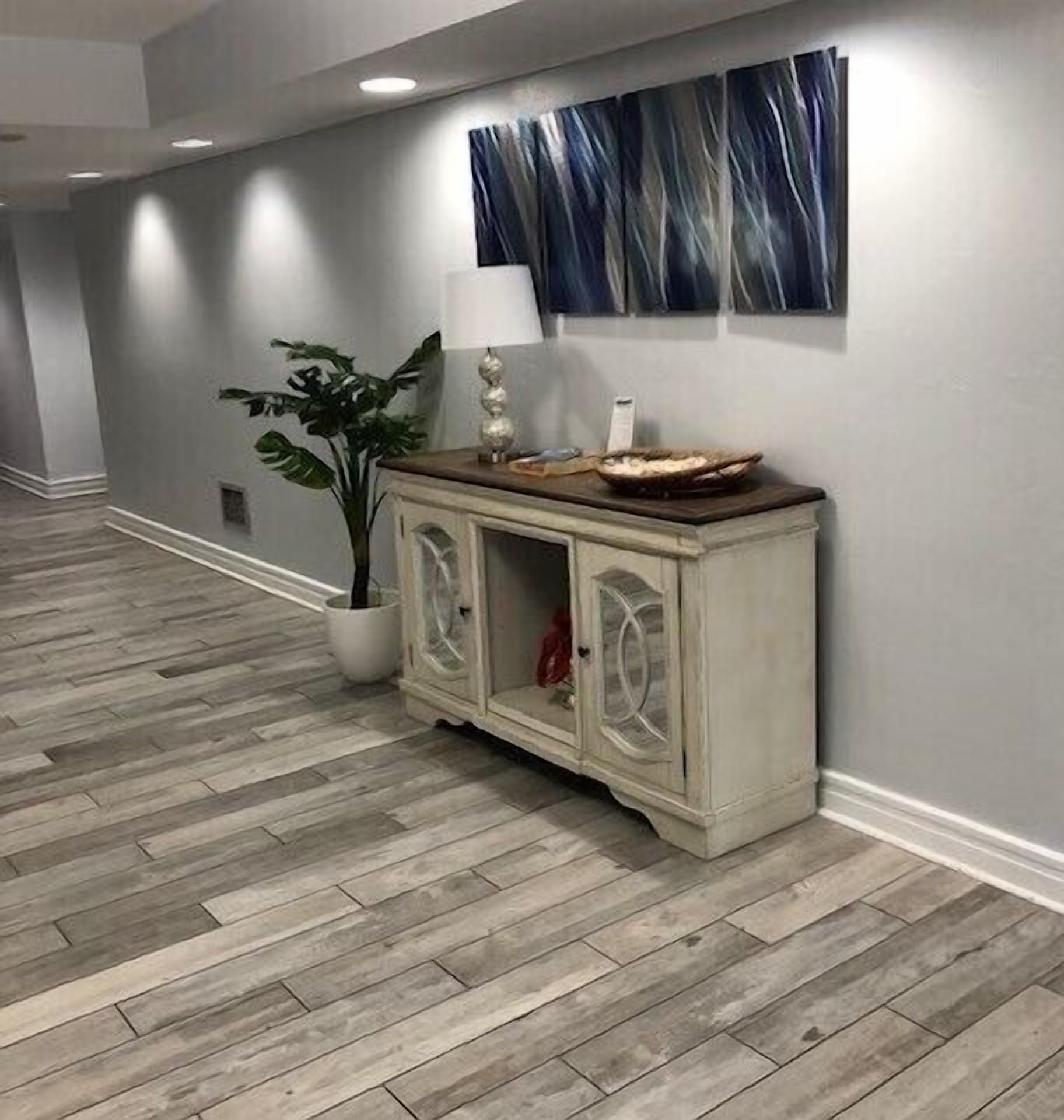 A hallway with a wooden floor, a white cabinet with glass doors, a potted plant, and a blue abstract painting on the wall.