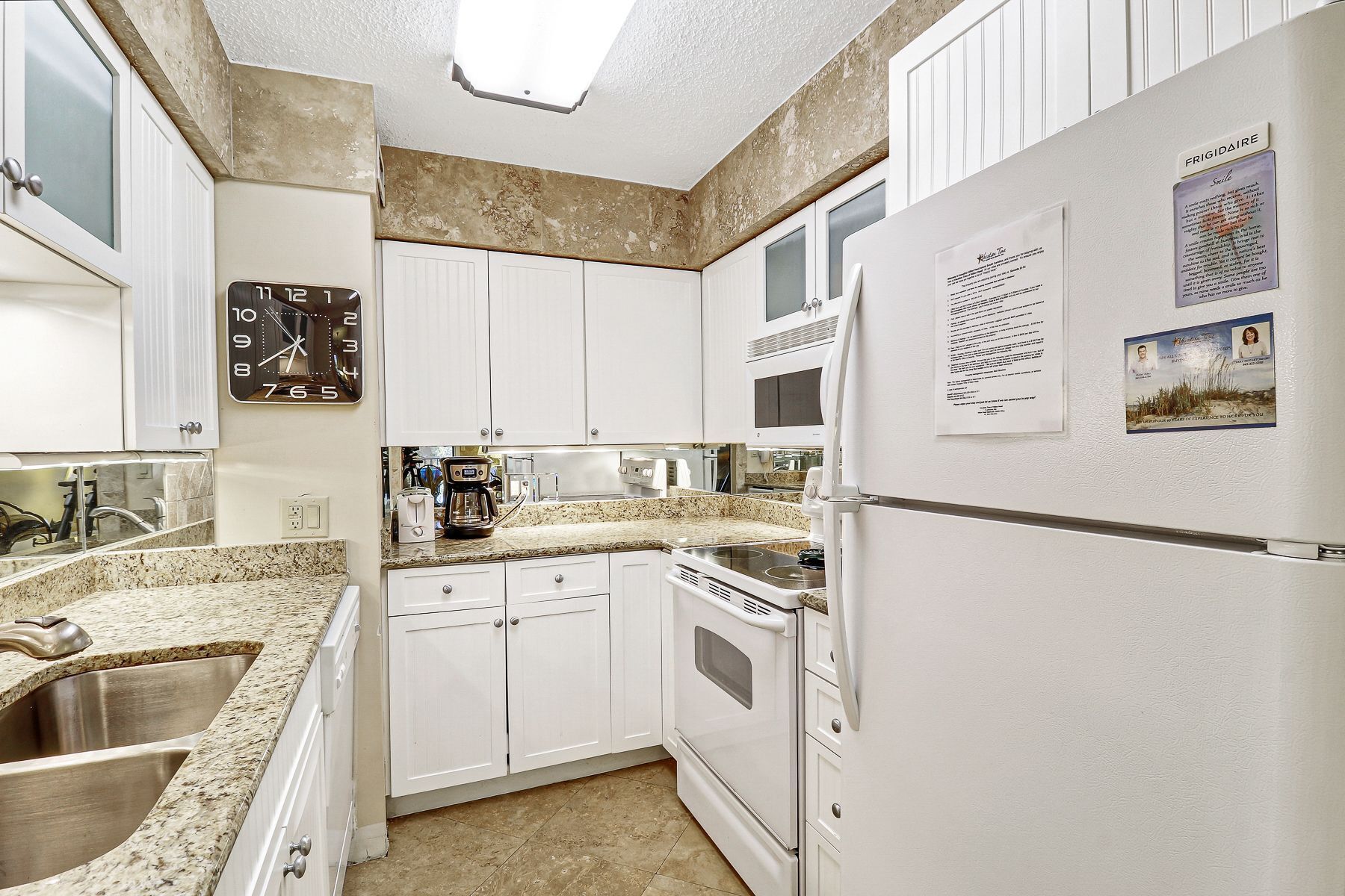 A kitchen with white cabinets, a stainless steel sink, a built-in oven, and a refrigerator. There is a clock on the wall and a menu on the refrigerator door.