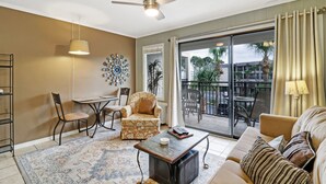 Un salón espacioso con sofá, sillón y una mesita. Hay un balcón con vistas a palmeras y edificios.