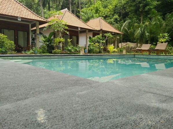 Uma piscina com deck ladrilhado, rodeada por vegetação exuberante e dois edifícios tipo bungalow com telhados em cerâmica.